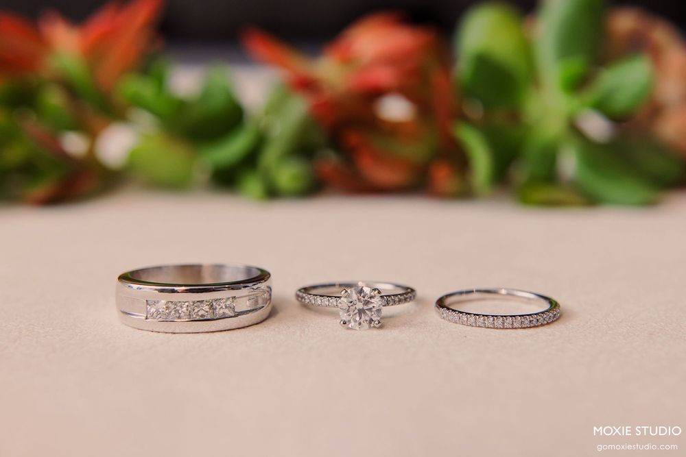 Three wedding rings including a diamond engagement ring and wedding bands displayed on a pink surface with floral decor.