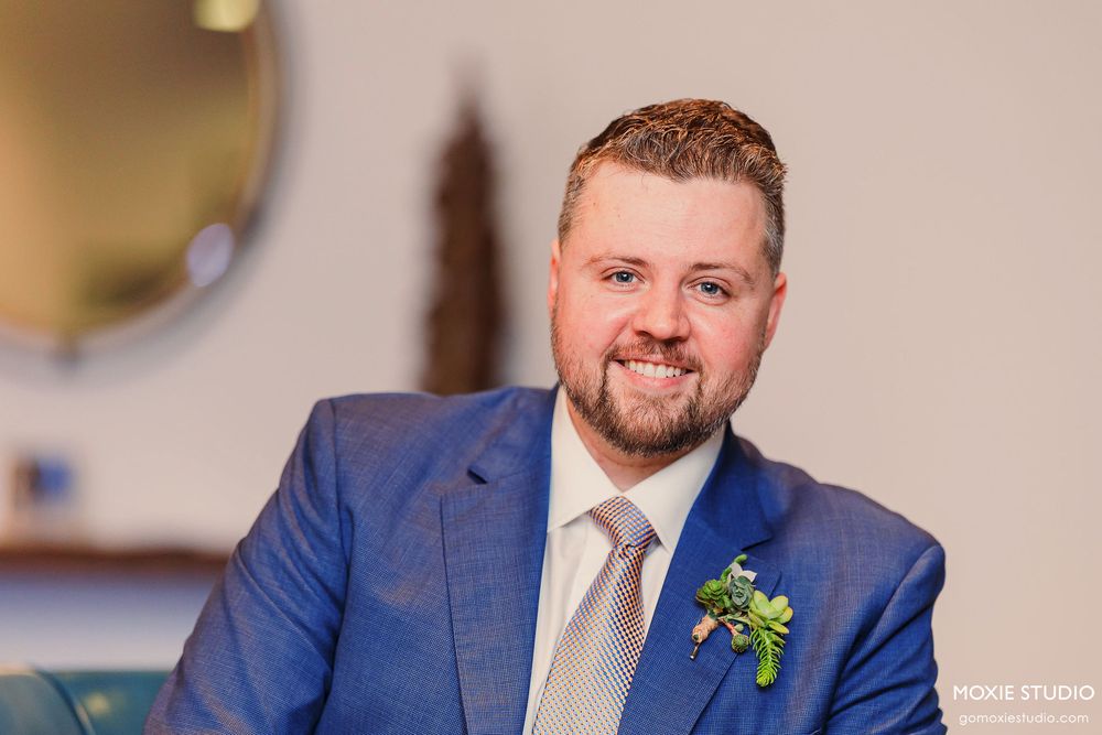 A person in a blue suit and beige tie smiles while standing in a formal setting.
