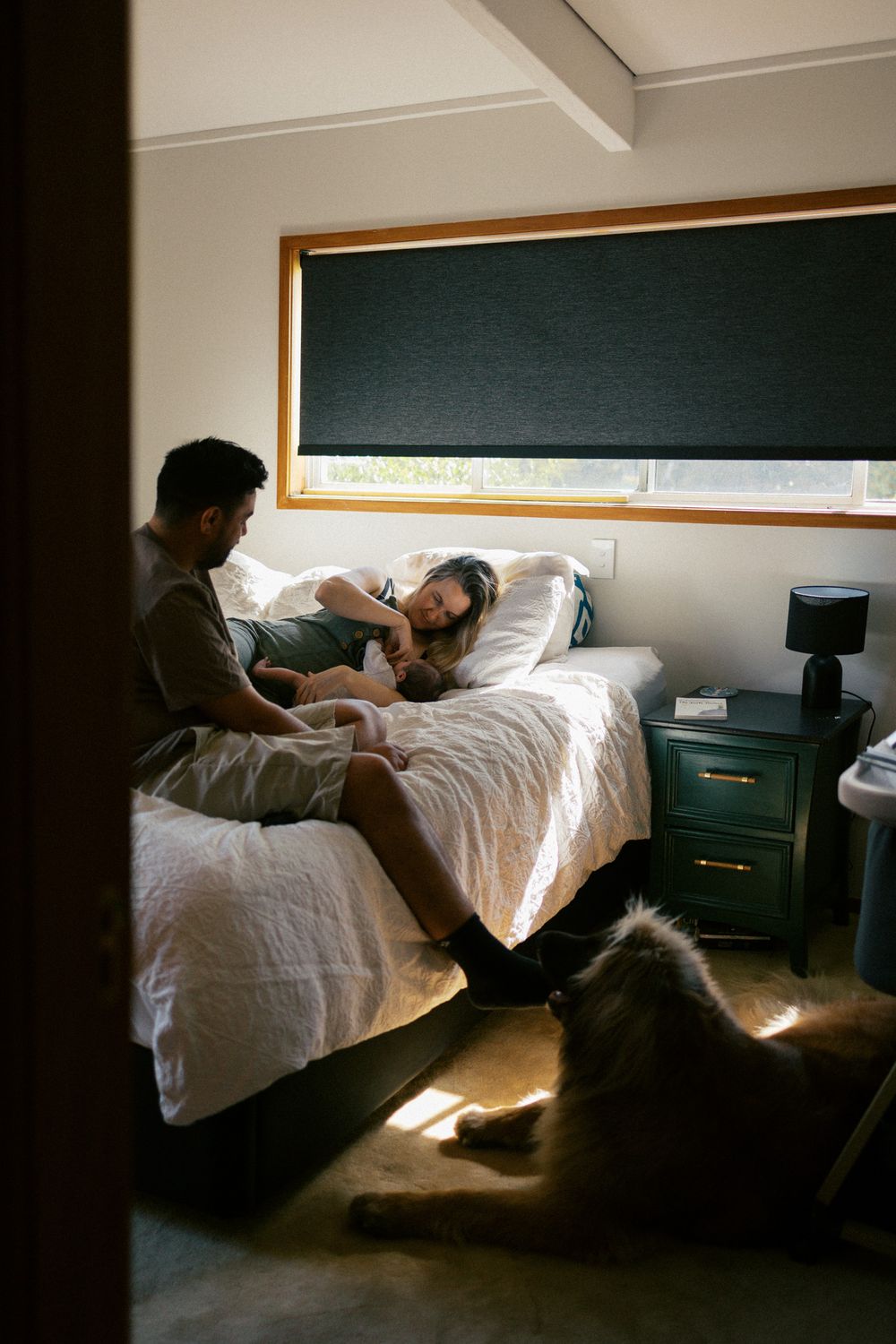 Intimate bedroom scene with couple relaxing on bed while dog rests nearby in natural window light.