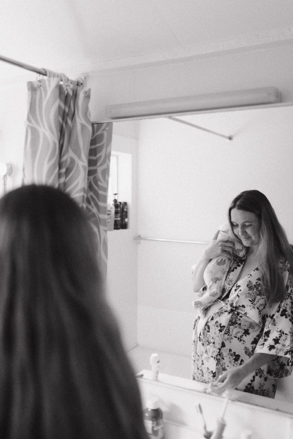 A black and white reflection in a bathroom mirror shows someone wearing a floral dress while looking down.
