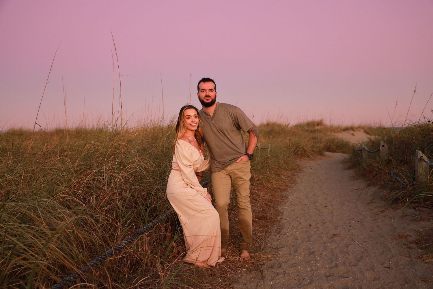 Couple embraces on sandy beach path surrounded by sea grass during pink sunset.