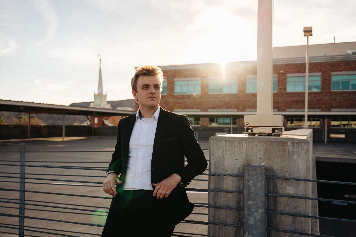 Man in business attire leans against railing on urban rooftop during golden hour sunset.