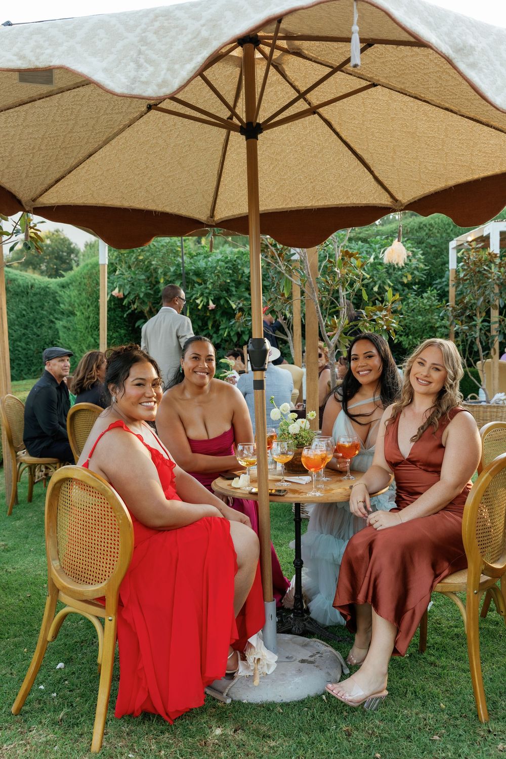 Wedding guests in red dresses relax under beige umbrellas during an outdoor reception.