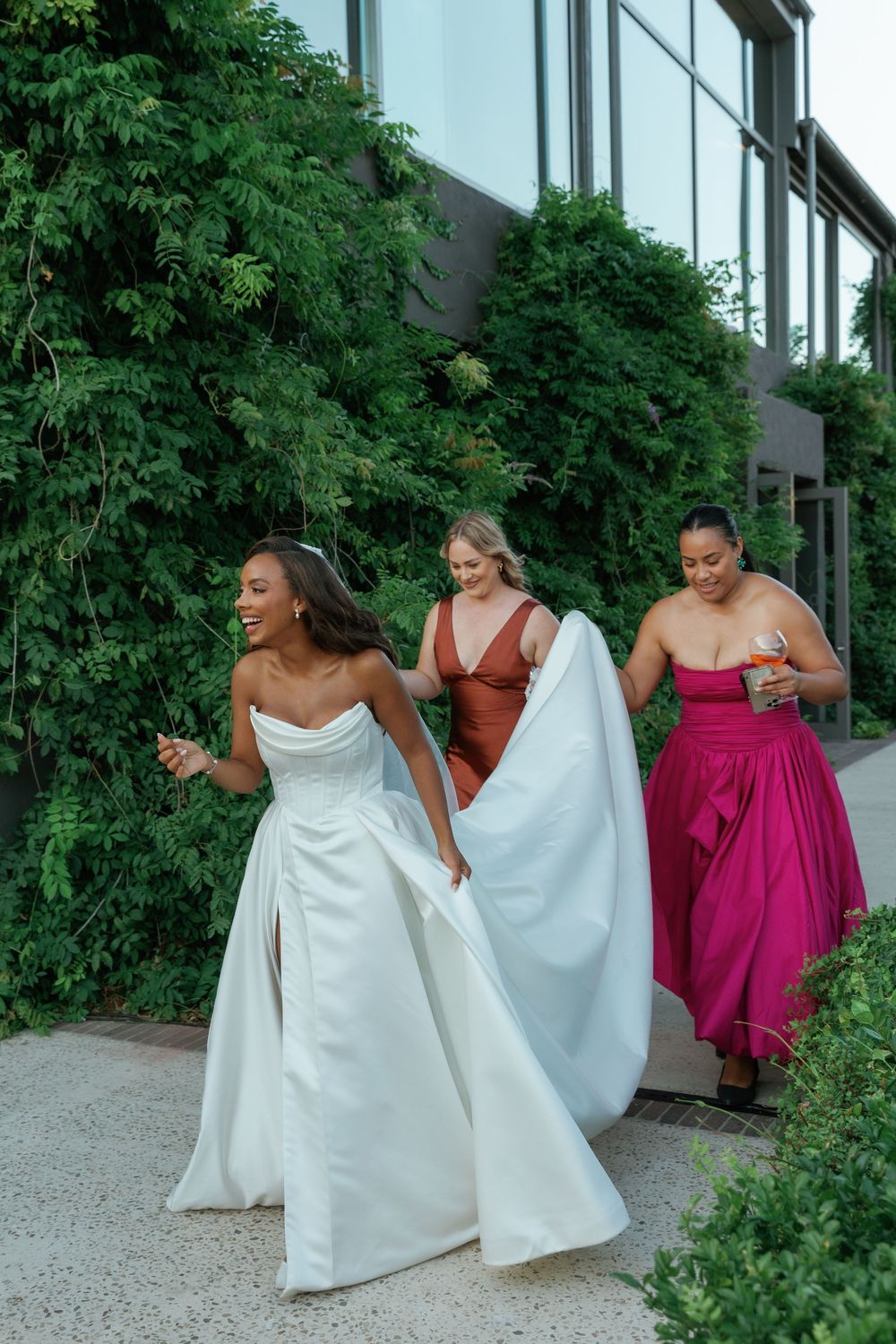 Wedding party in colorful dresses posing playfully outside a building with green foliage.
