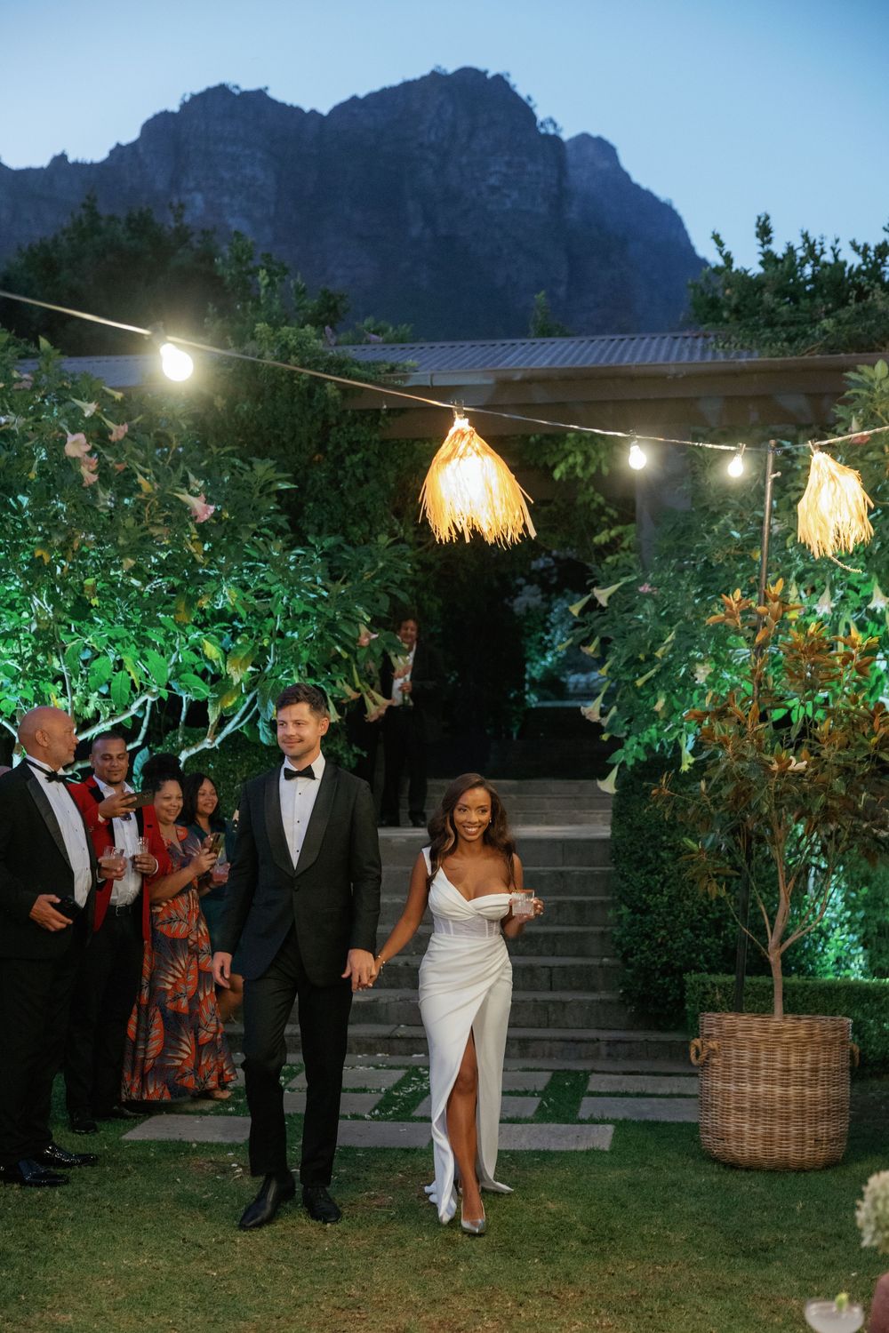 An evening wedding celebration with string lights and mountain backdrop as guests gather outdoors.