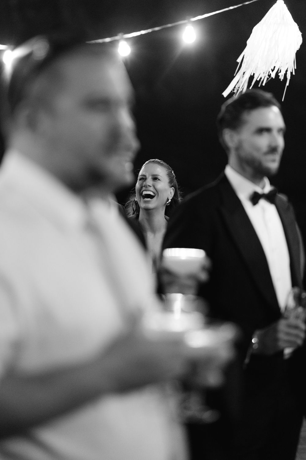 A candid black and white moment at an elegant party with string lights overhead showing joyful laughter.