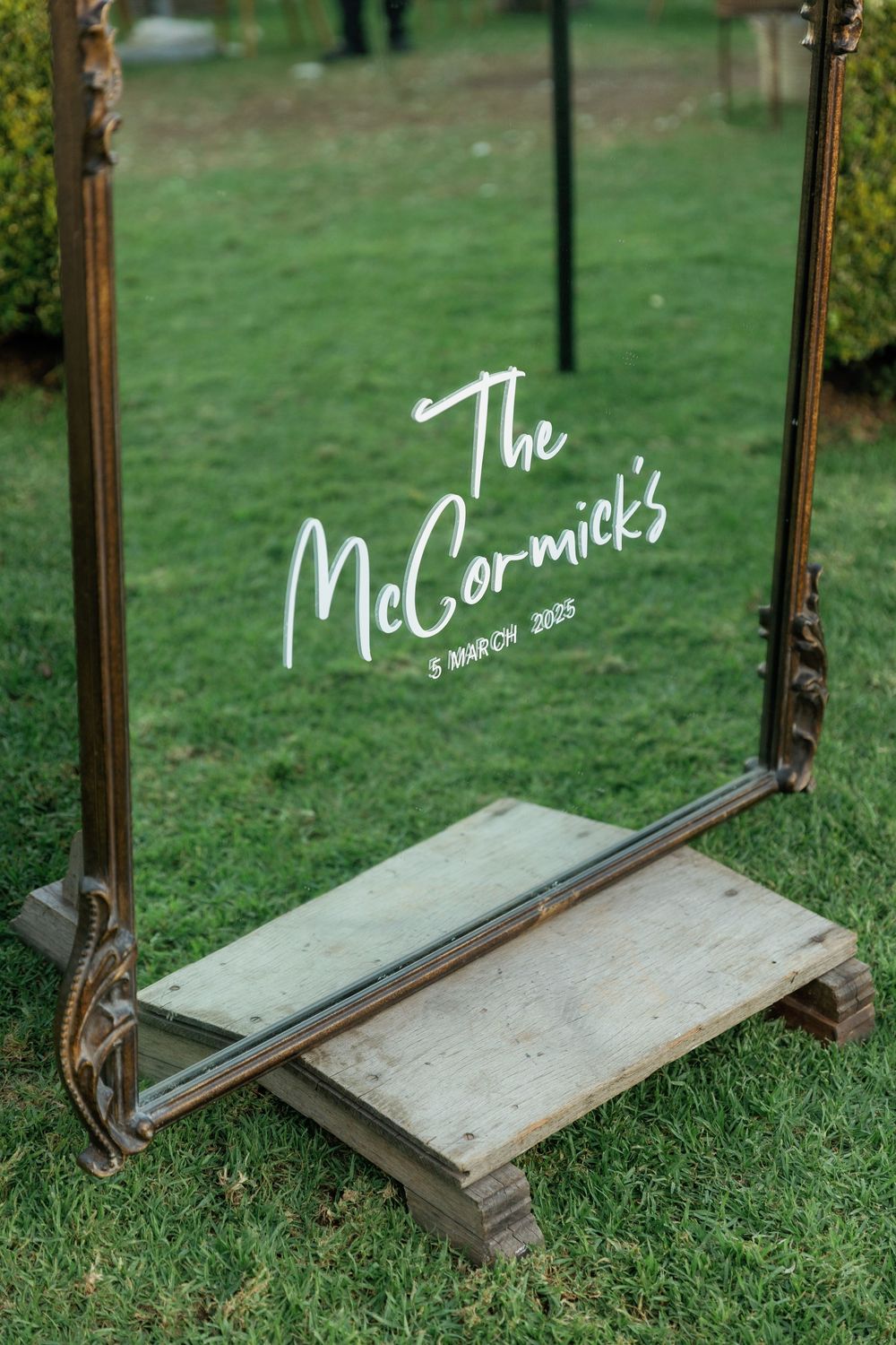Rustic wooden mirror frame with white text writing displayed on grass in an outdoor setting.