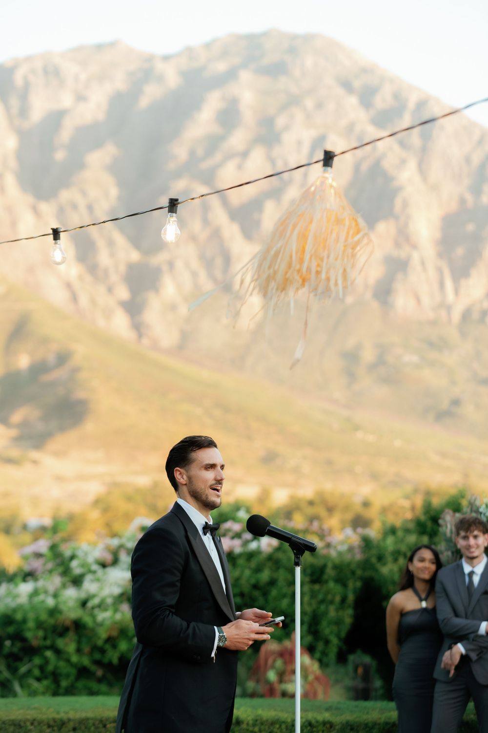 A wedding speech being given outdoors against a backdrop of majestic mountains with string lights overhead.