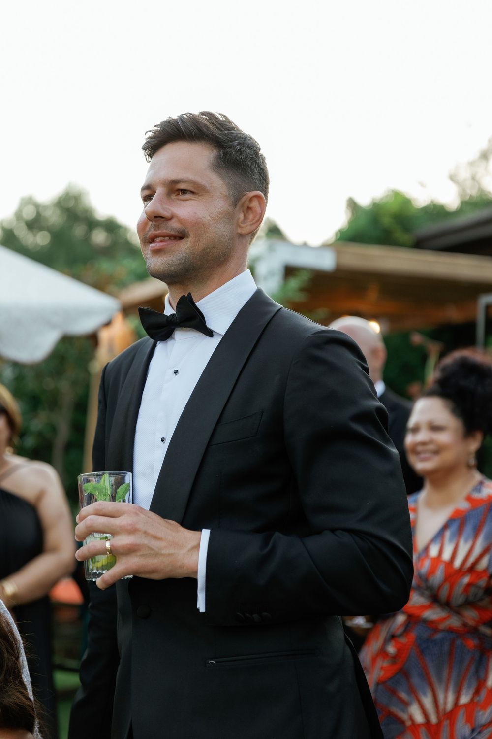 A guest in a black tuxedo and bow tie holds a drink at an outdoor wedding reception.