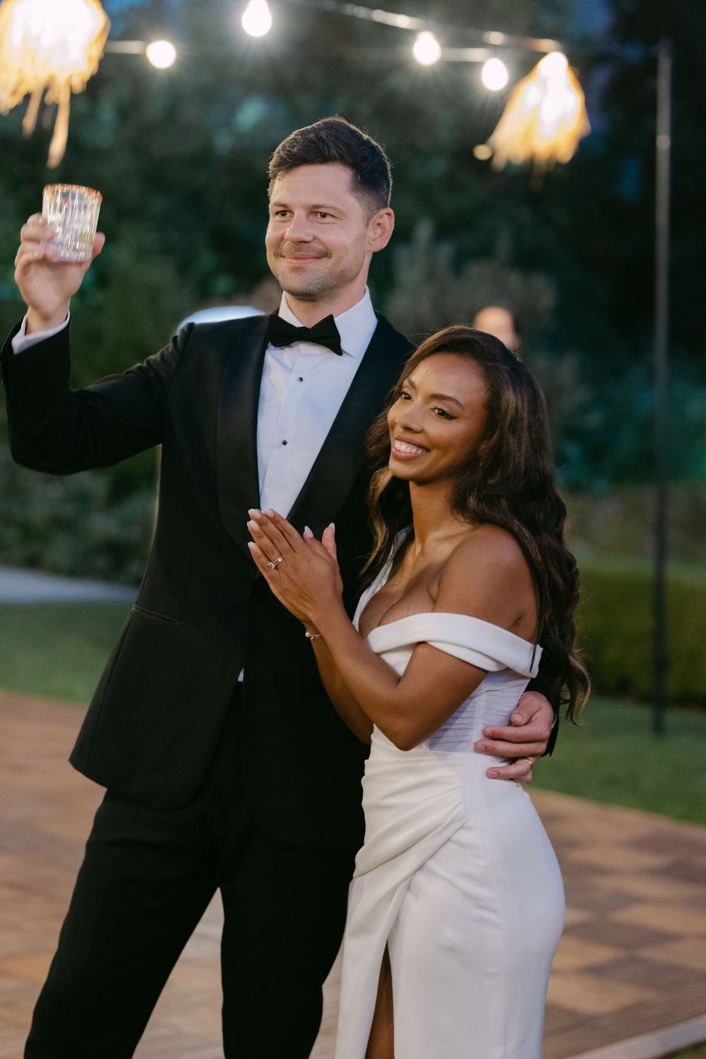 A joyful couple in formal attire share a romantic dance moment outdoors at dusk with string lights glowing above.
