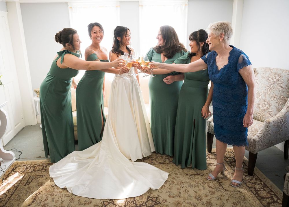 Bridesmaids in green dresses help adjust the bride's white wedding gown in a bright indoor setting.
