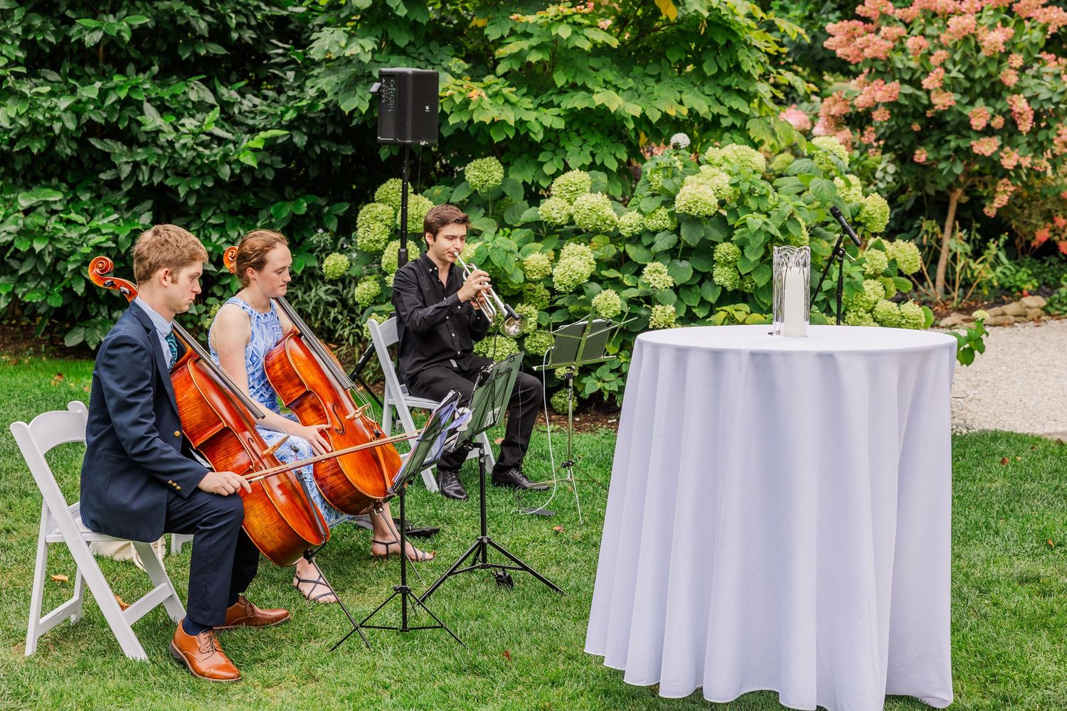 String musicians perform outdoors at an elegant garden event with white tables and lush greenery.