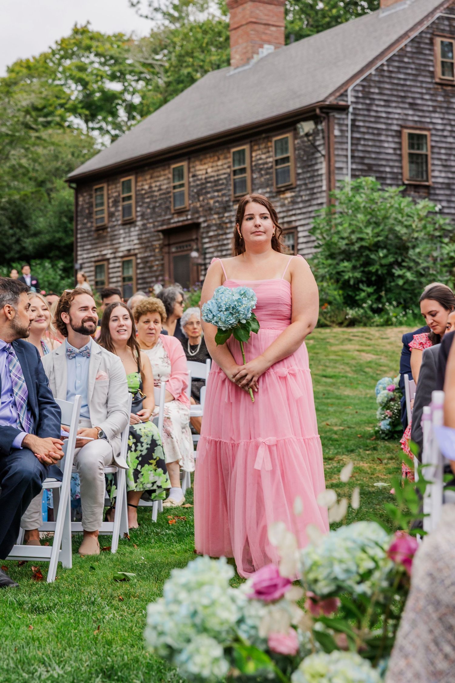 An outdoor wedding ceremony at a rustic colonial-style house with guests seated on white chairs and floral decorations.
