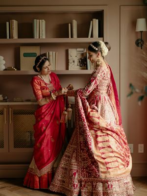 Two people in traditional Indian wedding attire with intricate red lehengas share a moment in front of built-in bookshelves.