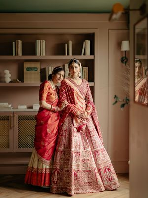 Two women in traditional Indian attire pose together in an elegant room with built-in shelving and soft lighting.
