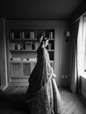 A person in an ornate embroidered lehenga stands by a window with built-in shelving in black and white photography.