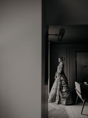 A black and white photograph shows a woman in a vintage Victorian-style dress standing in a dark room.