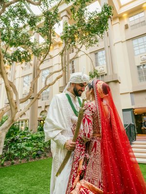 A traditional South Asian wedding couple in elegant attire poses under trees in a courtyard setting.