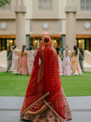 A red and gold Indian wedding lehenga dress displayed in multiple angles on a lawn with people in pink dresses in background.
