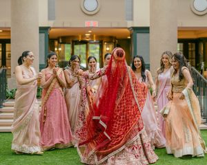 A group of women in pink and red lehengas and traditional Indian wedding attire pose together on a lawn.