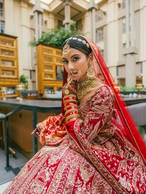 Traditional Indian bride in red embroidered lehenga poses elegantly against modern apartment building backdrop.