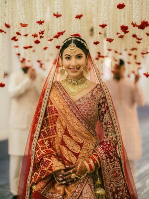 Traditional Indian bride in red lehenga under floral decor with hanging red roses.