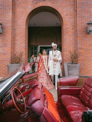 A wedding couple in traditional South Asian attire poses by a vintage red convertible car near brick archway.