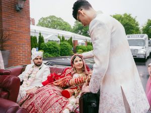 Traditional South Asian wedding ceremony participants in formal attire gather near a red car outside a brick building.