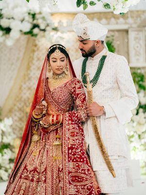 Traditional South Asian wedding couple in ornate red and white attire posing during their ceremony celebration.