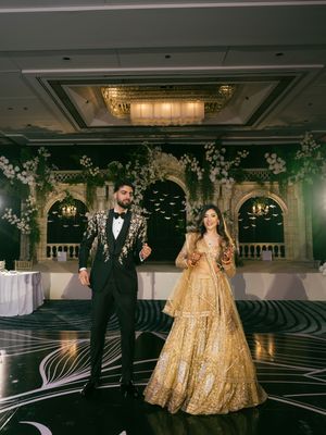 A couple in formal attire poses in an elegant ballroom with ornate decor and crystal chandeliers.