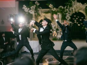 Wedding party members in black tuxedos perform synchronized dance moves at an elegant indoor reception.