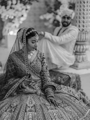 A bride in an ornate embroidered red lehenga sits gracefully in a garden setting during a traditional Indian wedding photoshoot.