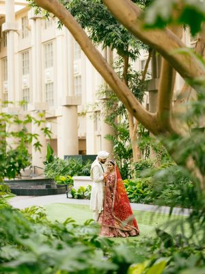 A couple in traditional wedding attire stands together among lush greenery outside an elegant building.