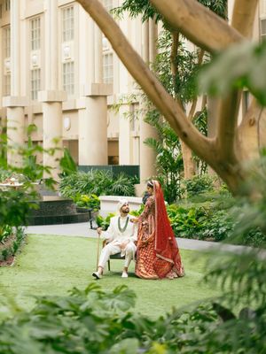 Couple in traditional Indian wedding attire poses on a manicured lawn outside a grand colonial-style building.
