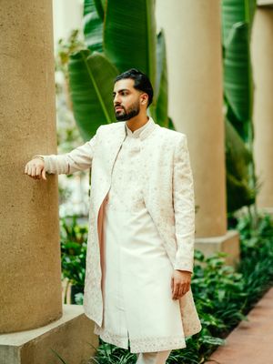 A person wearing a white embroidered sherwani poses elegantly against stone pillars in an outdoor setting.