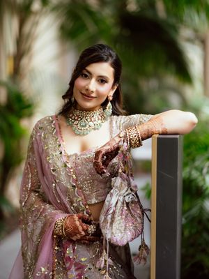 A bride in an embellished dusty rose lehenga poses elegantly against a natural outdoor backdrop.