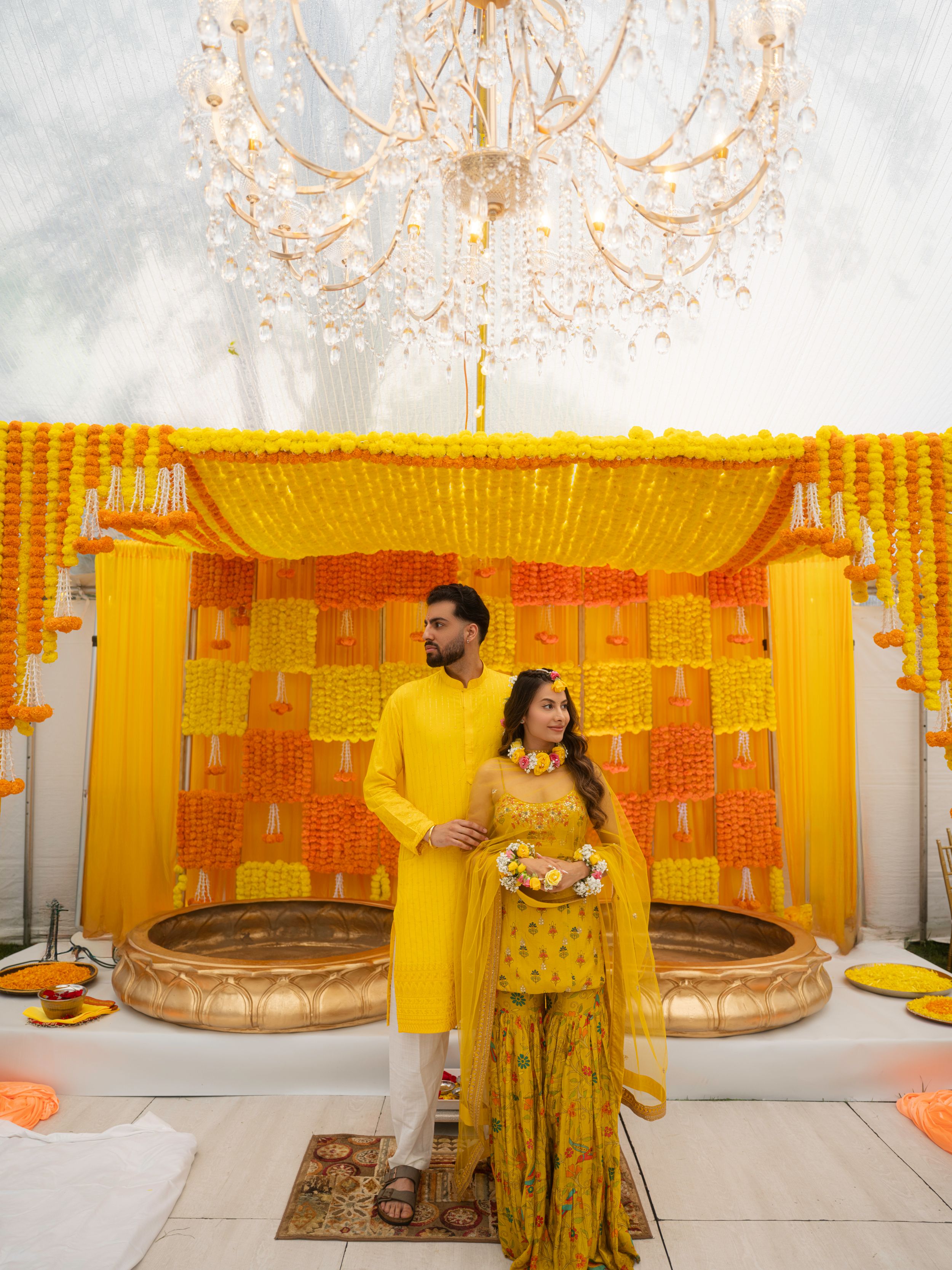 Traditional Indian wedding decor features vibrant orange and yellow marigold garlands with elegant white chandelier lighting.