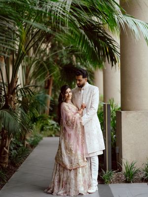 A couple in traditional wedding attire share romantic moments under a covered walkway with palm trees and columns.