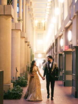 A couple in formal attire walks through an elegant colonnade hallway with soft natural lighting.