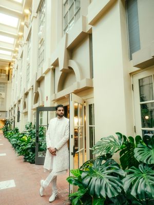 Person in white traditional attire walks along a corridor with lush monstera plants and architectural details.