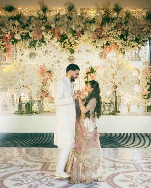 A couple in traditional South Asian wedding attire dance together under an elegant floral arch with pink and white blooms.