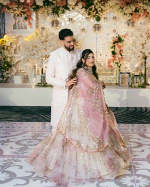 A couple in elegant wedding attire dancing and twirling on a patterned marble floor with floral decor in the background.