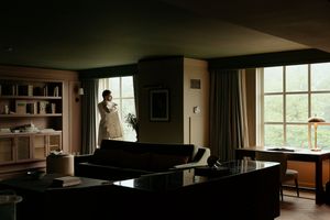 A silhouetted figure stands by large windows in a moody living room with dark furniture and natural lighting.