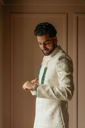 A groom in an elegant white embroidered sherwani smiles while adjusting his watch in a warm-lit room.