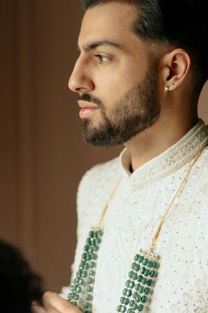 A close-up profile view of traditional Indian groom attire featuring an embroidered white sherwani and emerald necklace.
