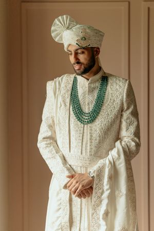Groom wearing traditional white embroidered sherwani with emerald green beaded necklace and matching turban for Indian wedding.