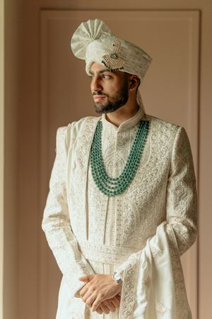 Traditional Indian groom wearing white sherwani with intricate embroidery and layered emerald green necklace.