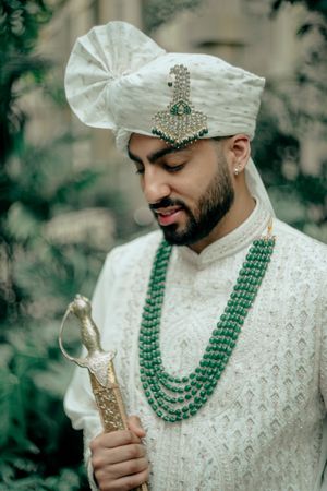 A groom in traditional Indian attire wearing an elegant white sherwani and emerald green beaded necklace during a wedding ceremony.