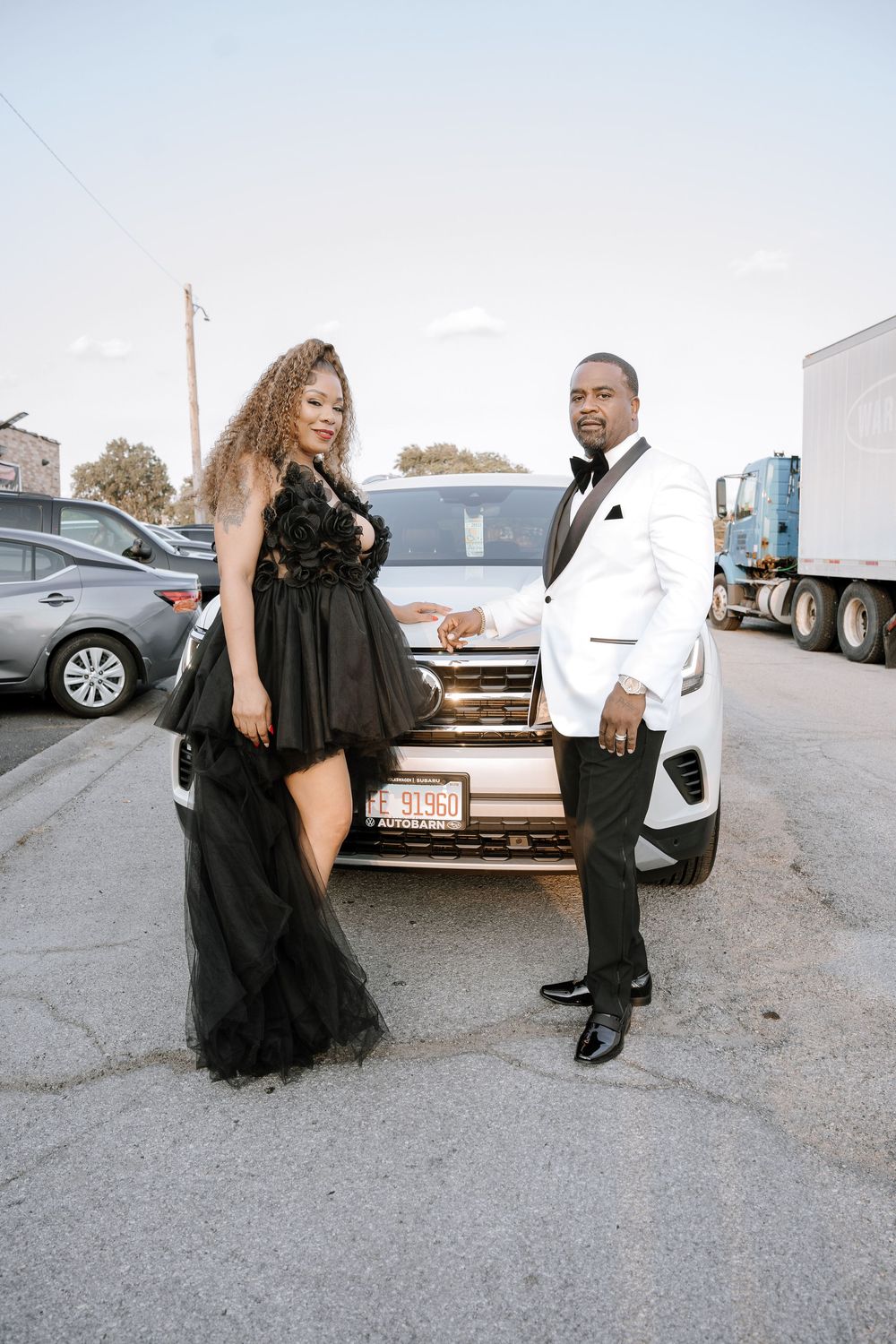 A couple shares a romantic moment in front of parked cars wearing black and white formal attire.