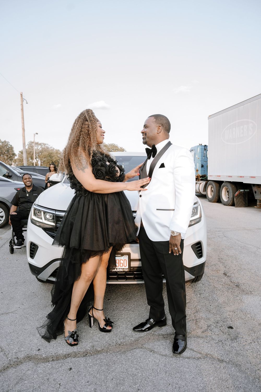 A couple in formal attire poses playfully in a parking lot with a white car in the background.